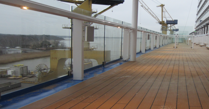 Laivan kannella on puinen lattia, lasikaiteet ja näkymä telakalle, jossa on nostureita ja vesistö.