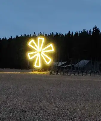Keltaisella neonvalolla valaistu tuulimylly seisoo pellolla metsän ja rakennusten edessä hämärässä.